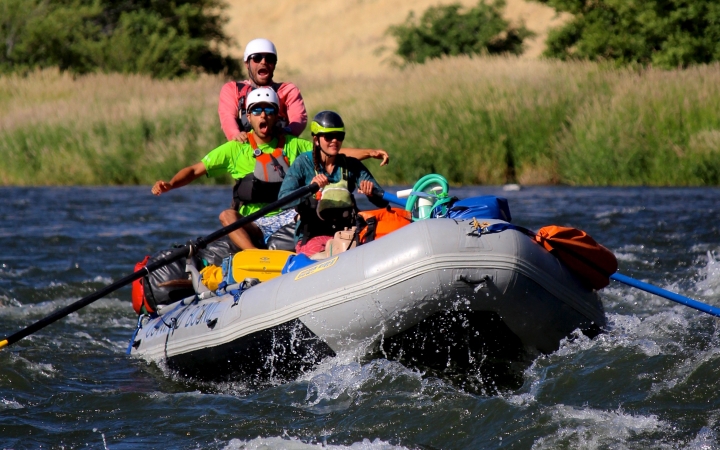 Three people wearing safety gear paddle a raft through whitewater.